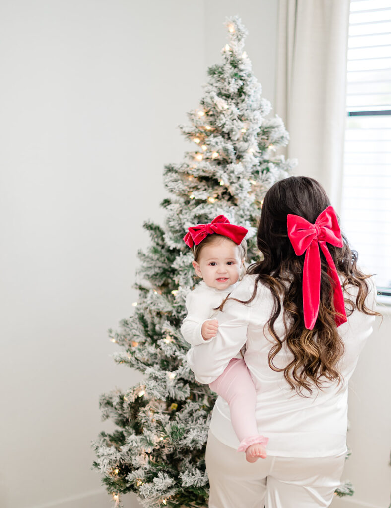 Mom and 1 year old baby matching Christmas pajamas and red hair bow in front of a christmas tree