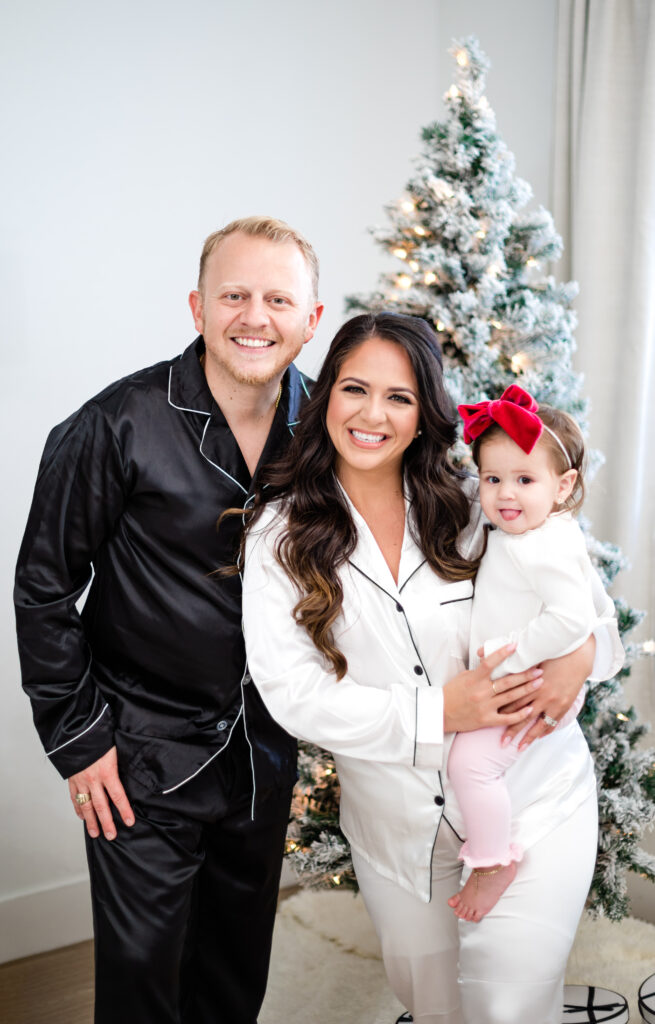 Mom and dad with a toddler matching Christmas pajamas in front of a christmas tree