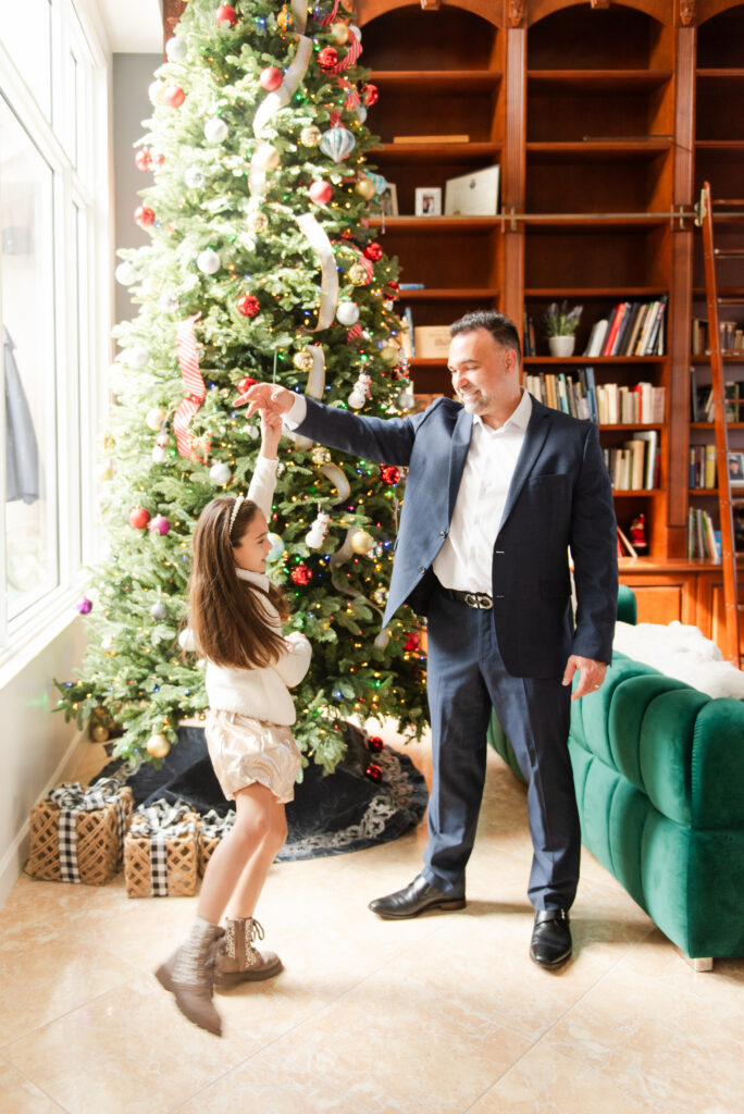 Father twirling daughter in front of the Christmas tree and bookcase at an in home for christmas photoshoot by MSP Lifestyle Miami Family Photographers